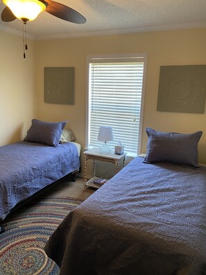 A room with two beds, a ceiling fan, a window with blinds, and a small table with a lamp.