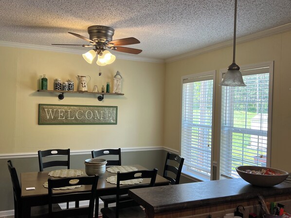 A dining area with a table set for four, a ceiling fan, and a 'WELCOME' sign on the wall.