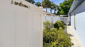 Et hvidt hegn med en skilt, der siger 'welcome', et overdækket udendørs siddeområde og en sti kantet med grøn bevoksning.
