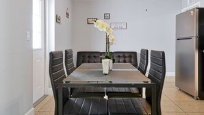 Et spiseområde med et sort bord og stole, en vase med hvide blomster og et køleskab i baggrunden.
