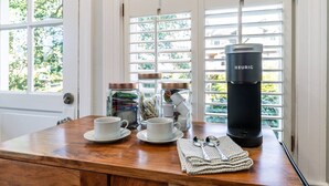 Une table en bois sur laquelle se trouvent deux tasses blanches, une machine à café Keurig noire, un bocal en verre contenant des sachets de thé et une paire de lunettes de soleil.