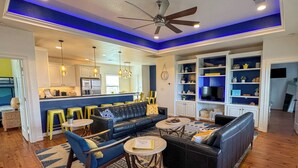 A modern living room with a blue accent wall, a black leather sofa, yellow stools, and a dining area with a kitchen in the background. - Family Beach House near Crystal Beach (Gilchrist) · Walk to the Gulf · Hot Tub (Gilchrist)