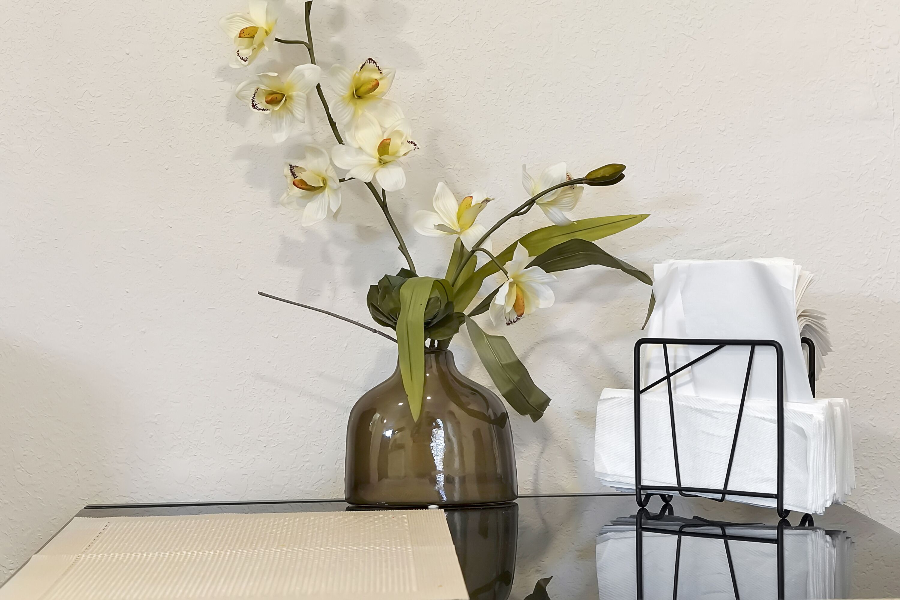 A glass table with a vase of white orchids, a black metal chair, and a stack of white napkins.