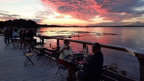 Vista del atardecer desde una terraza de madera con vista a un cuerpo de agua, con barcos, mesas y sillas para comer.