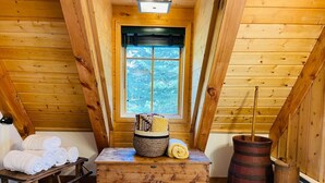 Eine Holzhütte mit Schlafzimmer im Zwischengeschoss, einer Holzbank mit Handtüchern, einem gewebten Korb und einem großen Holzbottich.