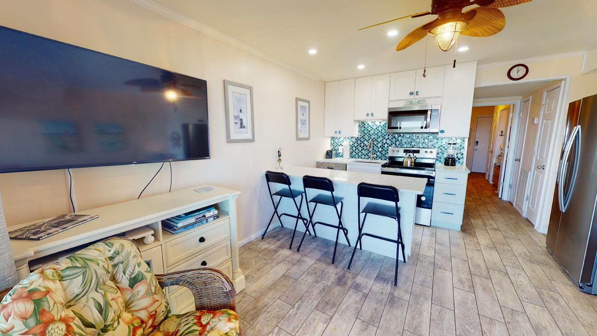 A modern kitchen with a central island, a dining area with bar stools, a flat-screen TV, and a ceiling fan.