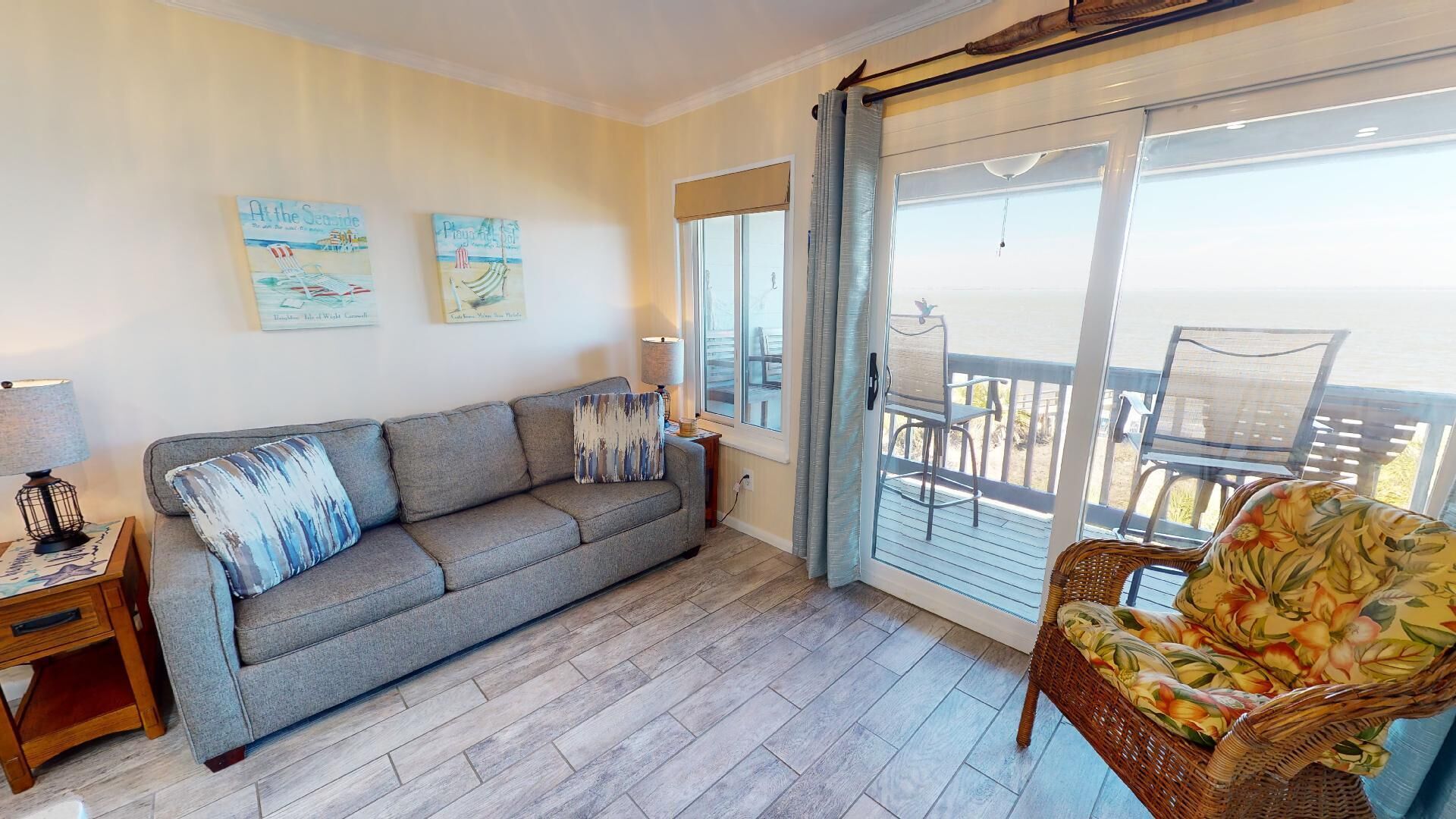 A living room with a grey sofa, a wicker chair, and a balcony with a view.