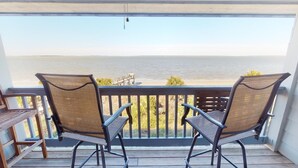 A balcony with two wicker chairs and a small table, overlooking a beach and a pier. - Coastal condo w. ocean views, community pool + beach access. (Tybee Island)