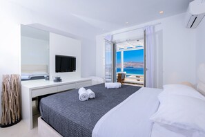 A modern hotel room with a large bed, a flat-screen TV, a desk, and a balcony with a view of the sea.