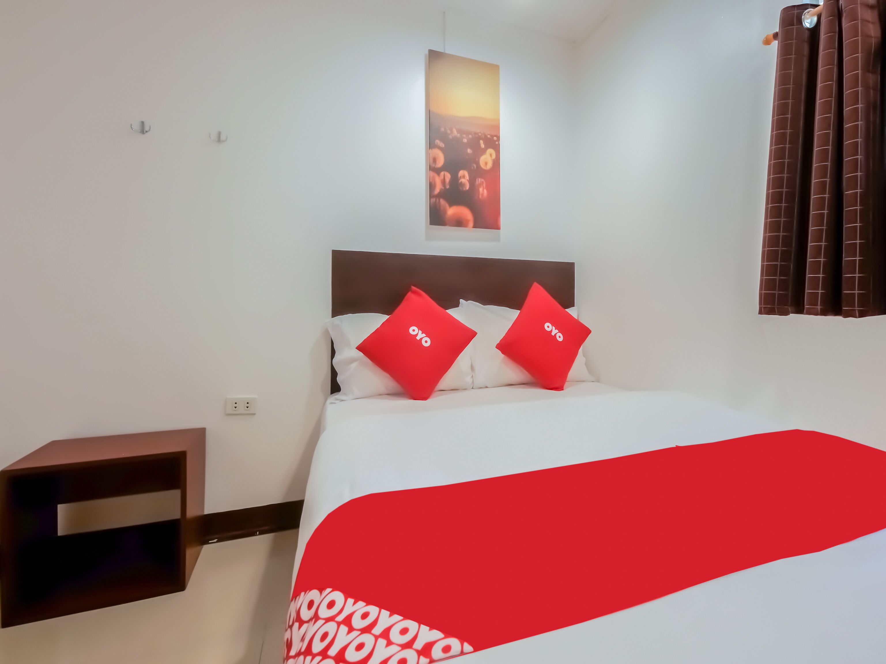 Una habitación de hotel con una cama, dos almohadas rojas, una colcha blanca, un cabecero marrón y una mesita de noche pequeña, además de una ventana con cortinas marrones.