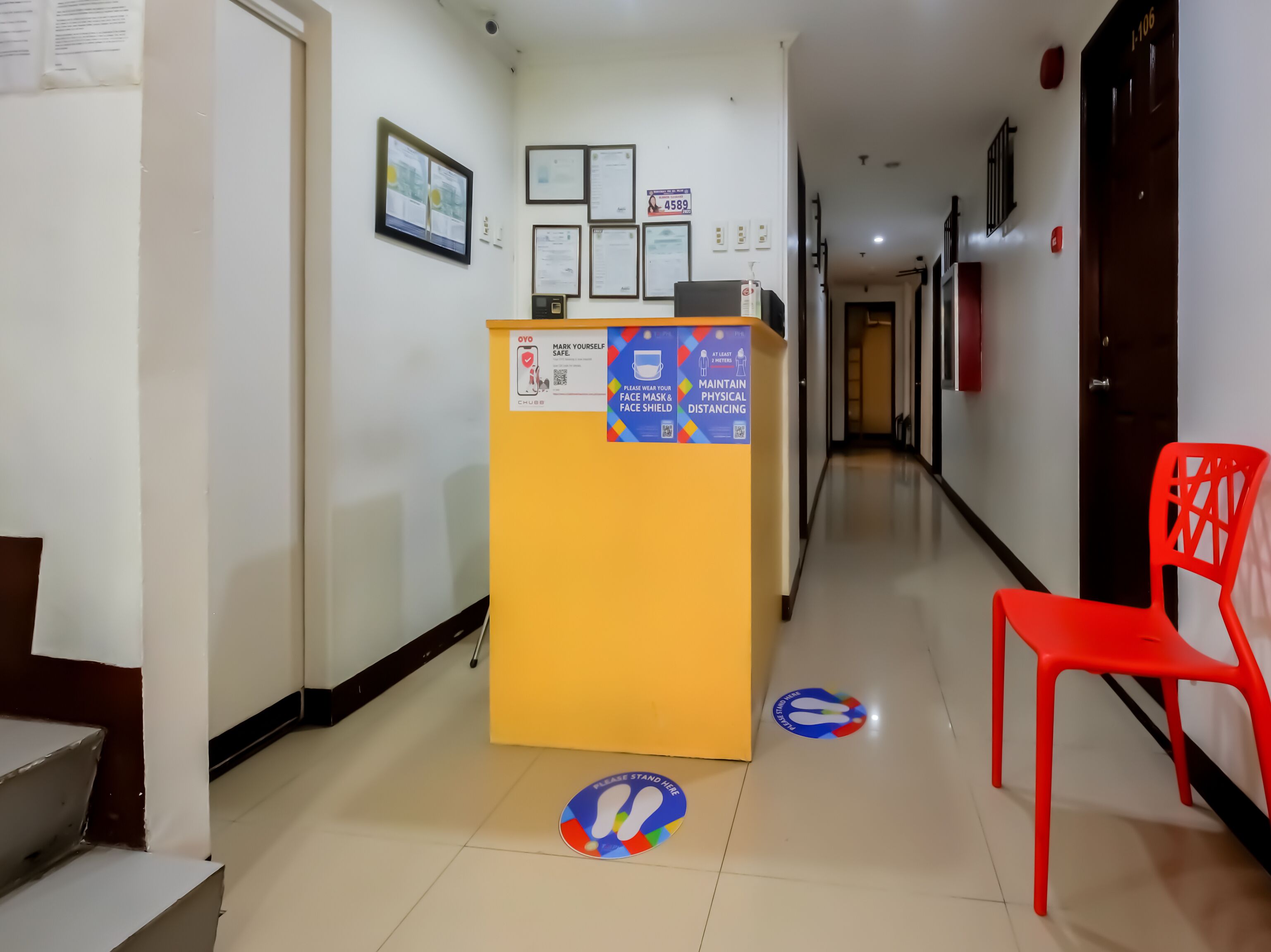 A hotel reception area with a yellow desk, red chairs, and floor markers for social distancing.