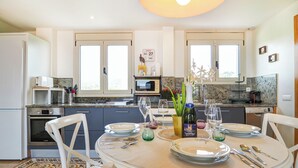 A modern kitchen with white cabinets, a dining table set for four, and a large window providing natural light. - Atico Los Condes by Hello Homes Sitges (Sitges)