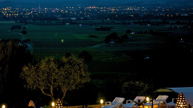 A terrace with lounge chairs and potted plants, overlooking a vineyard and a lit-up town in the distance.