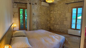 A bedroom with a stone wall, a wooden door, a bed with white bedding, and a window with a view of greenery.
