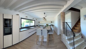 A modern kitchen with white cabinetry, a central island with bar stools, a stone column, and a staircase with a black railing.