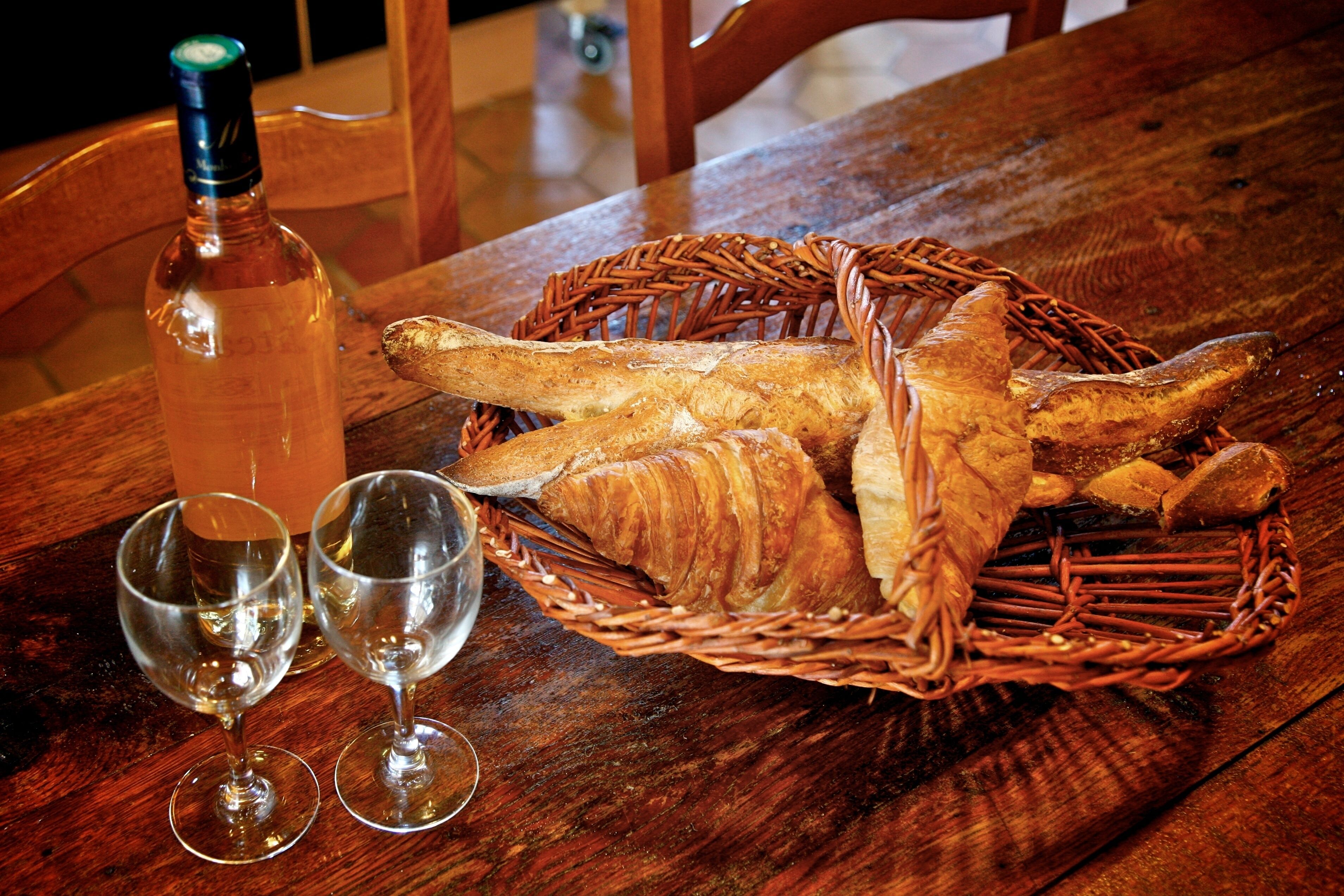 A wooden table with a bottle, two glasses, and a basket containing bread and croissants.