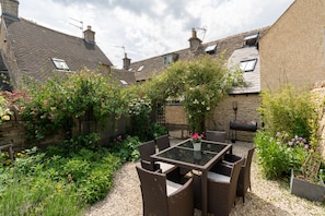 Une terrasse dotée d’une table en verre et de chaises en osier, entourée d’un jardin où poussent différentes plantes et fleurs.