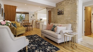 A living room with a sofa, armchairs, and a coffee table. There is a kitchen in the background with white cabinets and a stone wall. - The Staircase -  luxury dog friendly home in Stow on the Wold (Stow-on-the-Wold)