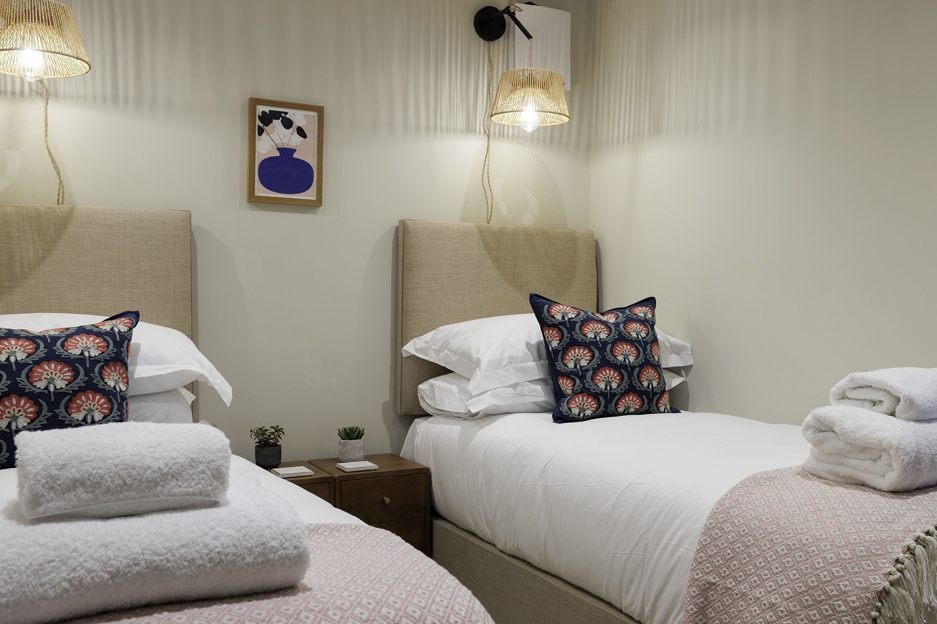 Habitación de hotel con dos camas, ropa de cama blanca y almohadas decorativas. Hay lámparas de pared y una obra de arte enmarcada sobre las camas.