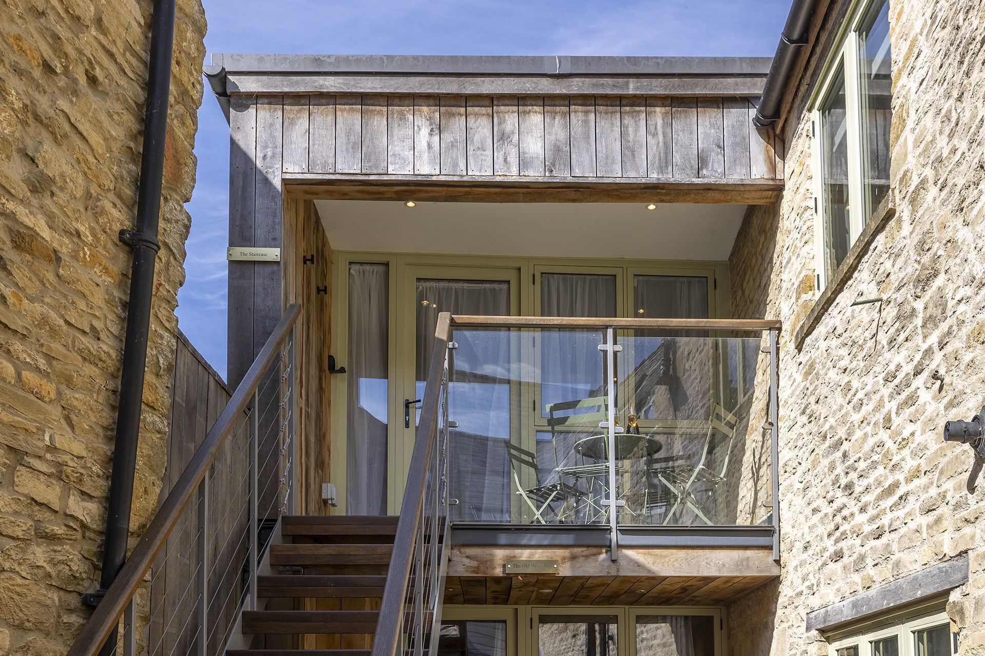A narrow staircase leading to a balcony with a glass railing, a stone wall on one side, and a door with a sign above it.
