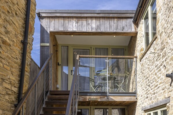 A narrow staircase leading to a balcony with a glass railing, a stone wall on one side, and a door with a sign above it.