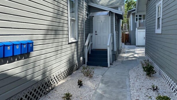 A narrow pathway leading to a white door, flanked by houses with grey siding and a blue mailbox on the left. - Serenity Bungalow Near Armature Works & Riverwalk (Tampa)