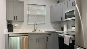A modern kitchen with grey cabinets, a stainless steel dishwasher, a sink, a stove, and a window with blinds. - Serenity Bungalow Near Armature Works & Riverwalk (Tampa)