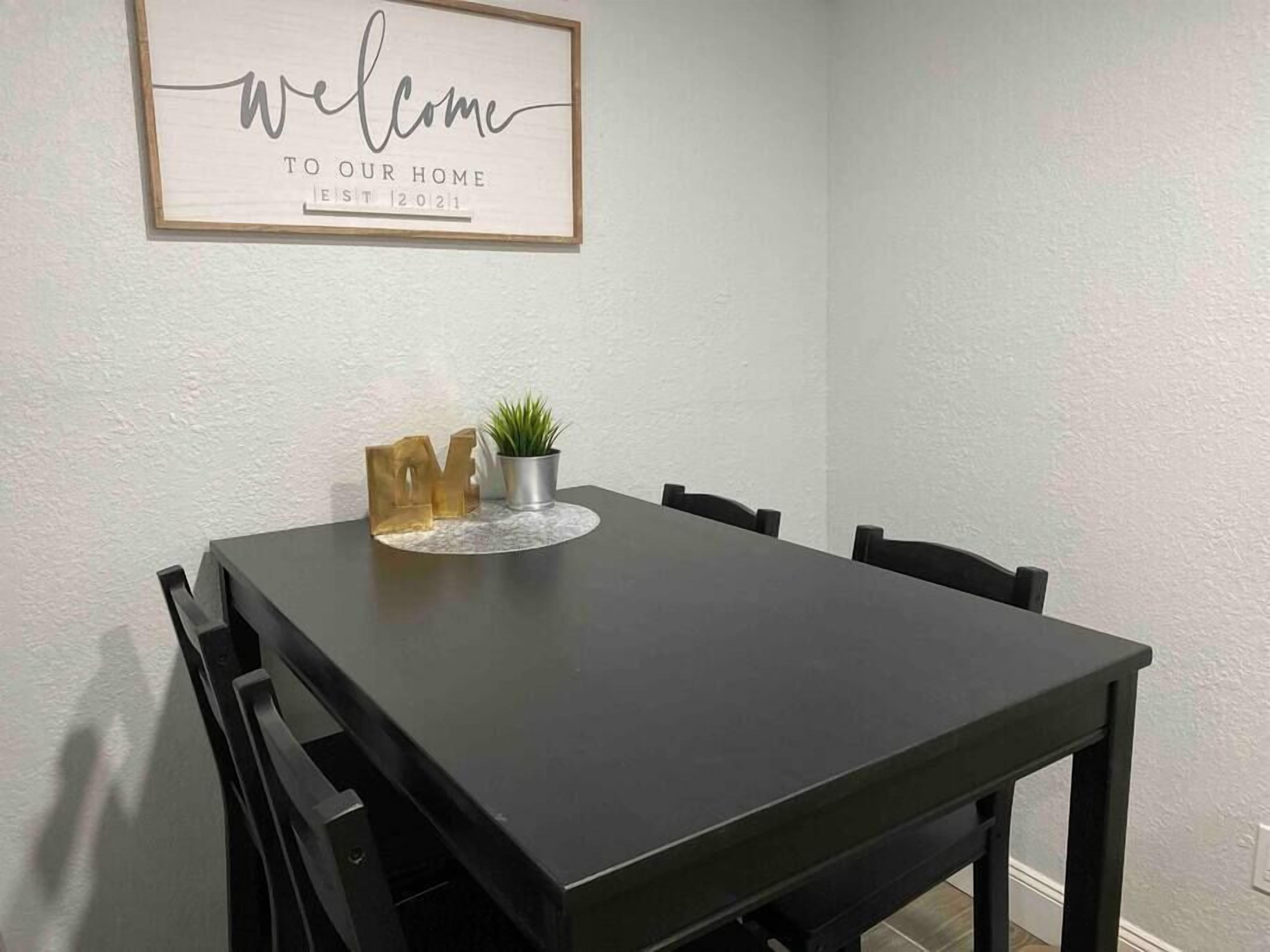 Una mesa de comedor con sillas, un cartel de bienvenida y una planta en maceta.