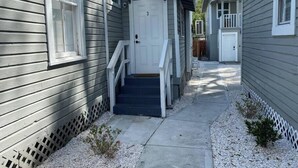 A narrow pathway leading to a white door, flanked by two-story buildings with grey siding and white trim.