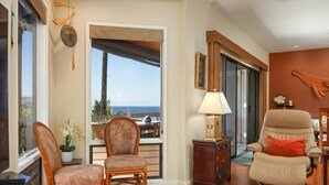 A living room with a view of the ocean, a wicker chair, a recliner, a lamp, and a wooden cabinet. - Aloha Condos, Kanaloa at Kona, Condo 3301, Ocean View, AC (Kailua Kona)