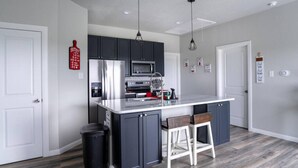 A modern kitchen with dark cabinets, a stainless steel refrigerator, a microwave, and a white countertop. There are two white bar stools and a black trash bin.