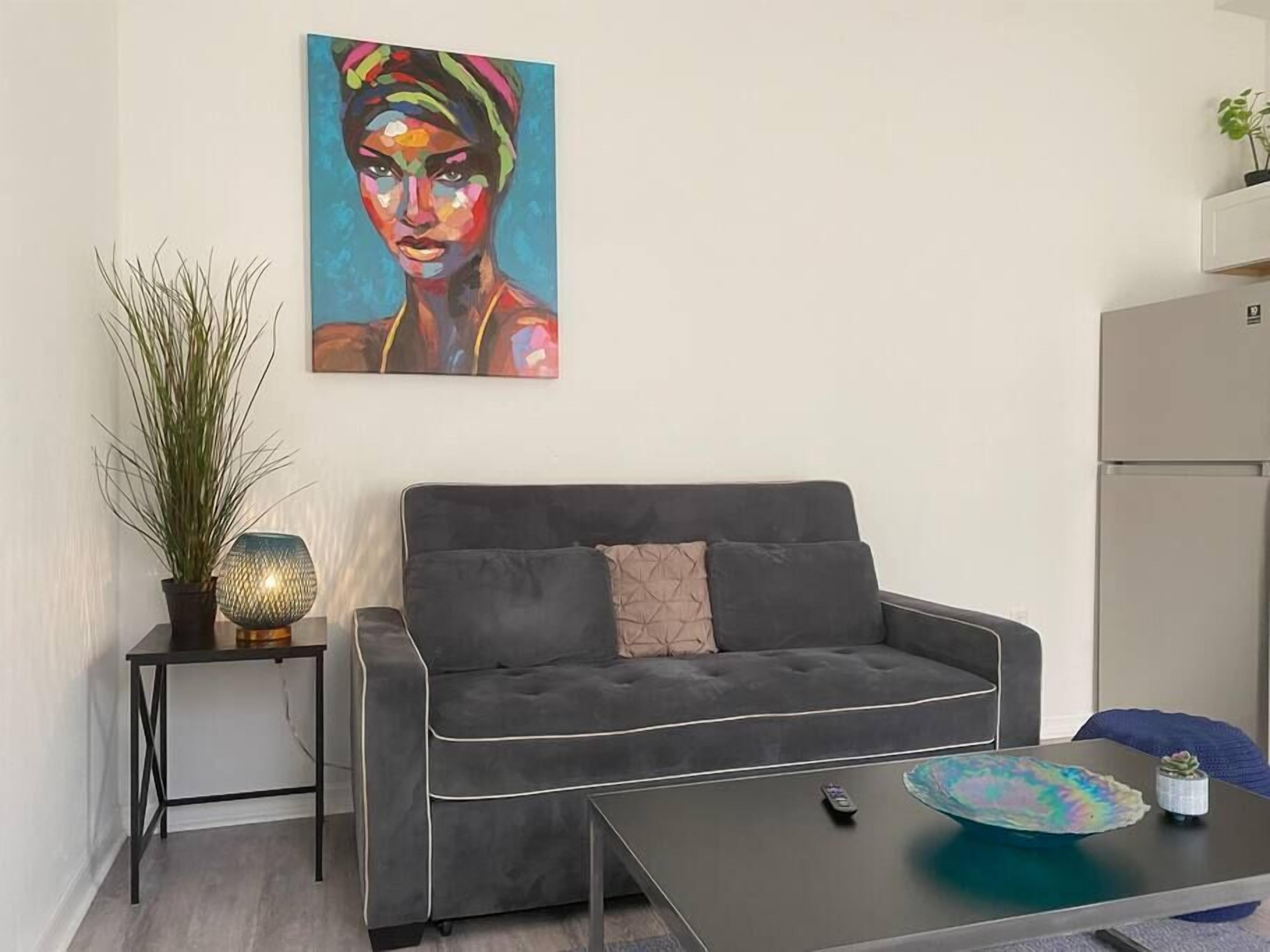 Een woonkamer met een grijze bank, een klein bijzettafeltje met een lamp, een schilderij aan de muur en een salontafel met een afstandsbediening.