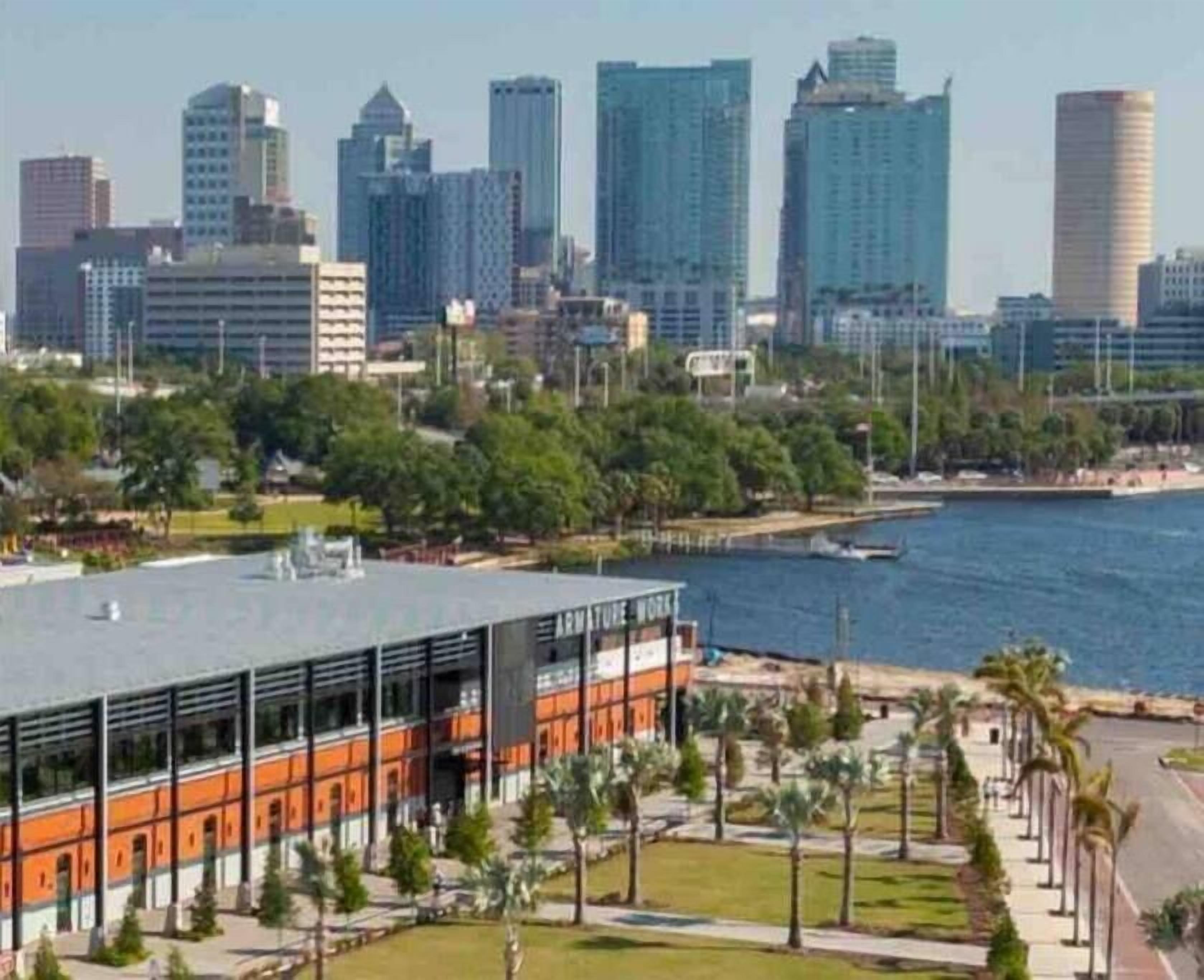 A modern building with a glass facade and orange accents, located near a waterfront with palm trees and a clear sky.