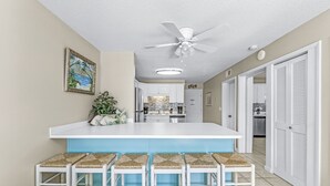 Une cuisine avec un îlot central blanc et des tabourets de bar, un ventilateur de plafond et un tableau encadré au mur.