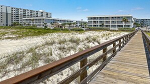 Une promenade en bois mène à un hôtel en bord de mer, doté de nombreux balcons et donnant sur une plage de sable.