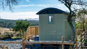 Eine kleine, eingeschossige Holzhütte mit geneigtem Dach, einem Fenster und einer Tür, umgeben von einer Holzterrasse und einem Zaun.