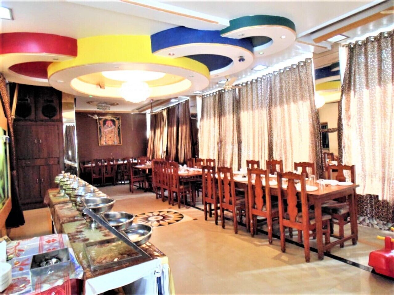 Un restaurante con varias mesas y sillas, una larga barra de buffet y un techo con lámparas circulares.