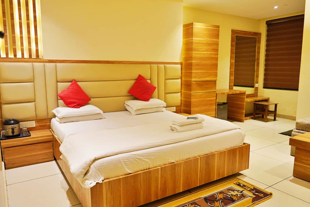 A hotel room with a large bed, two red pillows, a wooden headboard, a wooden nightstand, a small table, and a window with blinds.