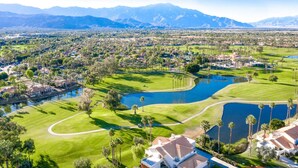 A golf course with a large pond, surrounded by residential areas and mountains in the background. - Near Coachella | Desert Falls Home | Steps to Pool, Spa, & Fun! Mountain Views Out Your Window! (Palm Desert)