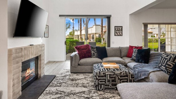 A spacious living room with a fireplace, a large flat-screen TV, and a view of a courtyard with palm trees. - Near Coachella | Desert Falls Home | Steps to Pool, Spa, & Fun! Mountain Views Out Your Window! (Palm Desert)