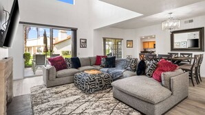 A spacious living room with a large sectional sofa, a flat-screen TV mounted on the wall, and a dining area with a chandelier. - Near Coachella | Desert Falls Home | Steps to Pool, Spa, & Fun! Mountain Views Out Your Window! (Palm Desert)