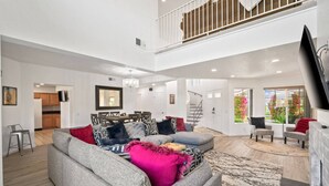 A spacious living room with a grey sectional sofa, a chandelier, and a dining area with a large mirror. - Near Coachella | Desert Falls Home | Steps to Pool, Spa, & Fun! Mountain Views Out Your Window! (Palm Desert)