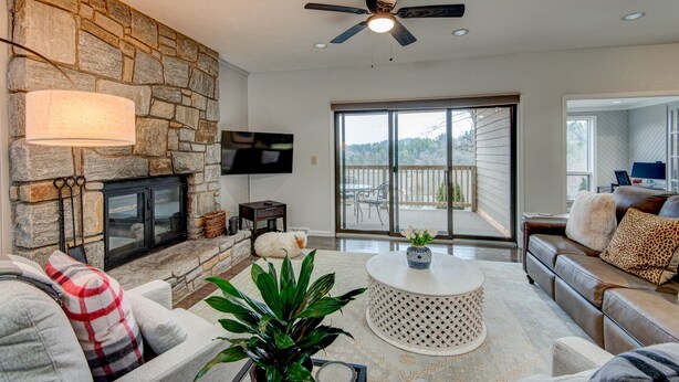 A modern living room with a stone fireplace, a ceiling fan, a flat-screen TV, and a balcony with a view. - Chetola Resort: Robin 1 - Lower Level Unit near downtown Blowing Rock (Blowing Rock)