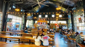 Un vaste espace intérieur aménagé en salle à manger, avec des bancs et des tables en bois, un plafond haut avec des poutres apparentes et des guirlandes lumineuses suspendues au plafond.