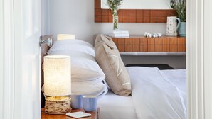 A neatly made bed with white linens, a woven bedside table with a lamp, and a wooden nightstand with a mirror. - Sea-la-vie, Astor House -  an apartment that sleeps 4 guests  in 1 bedroom (Torquay)
