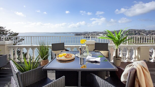 A terrace with wicker furniture, a table with breakfast items, and a view of the sea and distant landscape. - Sea-la-vie, Astor House -  an apartment that sleeps 4 guests  in 1 bedroom (Torquay)
