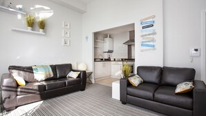 A modern living room with two leather sofas, a kitchen in the background, and a wall with nautical-themed signs. - Sea-la-vie, Astor House -  an apartment that sleeps 4 guests  in 1 bedroom (Torquay)
