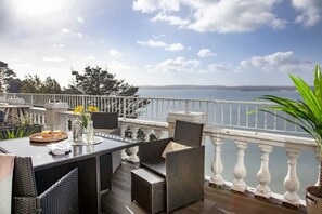 Eine Terrasse mit Korbmöbeln, einem Tisch mit einer Glasvase und einer Speisenauswahl, mit Blick auf das Meer.
