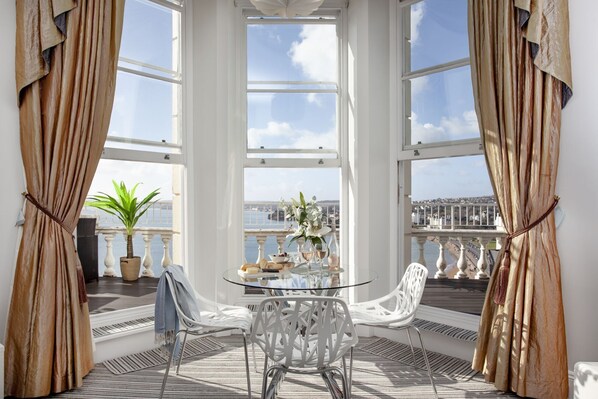 A sunlit room with a glass table, two white chairs, and a view of the sea through large windows.