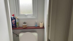 A small, compact bathroom with a white toilet, a window above the sink, and a wooden shelf with toiletries. - Mobile home for 6 people. (Arvert)